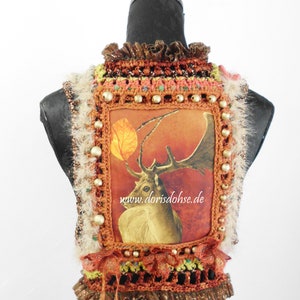 May include: A crocheted vest with a brown, orange, and green color scheme. The vest features a square panel with a printed image of a deer with antlers and a leaf. The panel is surrounded by crocheted trim and gold beads. The vest has a fringe trim at the bottom. The text "www.dorisdohse.de" is printed on the panel.