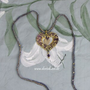 May include: A heart-shaped pendant necklace with a gold and purple design. The pendant is made of fabric and has a delicate, intricate pattern. The necklace is made of a thin, gray cord with gold accents. The pendant is hanging on a white background with a floral pattern.