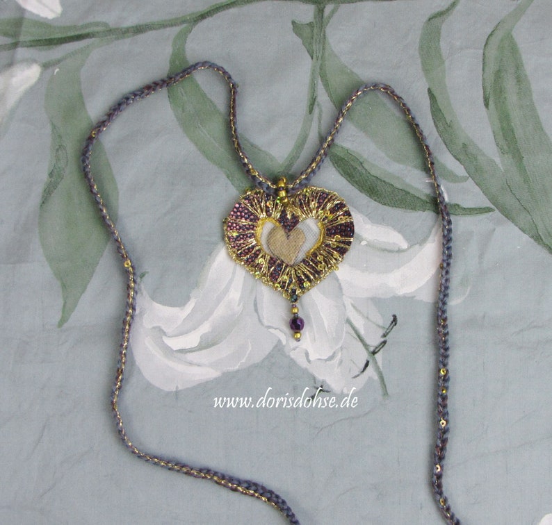 May include: A heart-shaped pendant necklace with a gold and purple design. The pendant is made of fabric and has a delicate, intricate pattern. The necklace is made of a thin, gray cord with gold accents. The pendant is hanging on a white background with a floral pattern.