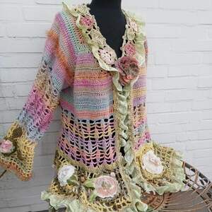May include: A multi-colored crochet cardigan with a floral design. The cardigan has a light green, pink, and yellow color scheme. The cardigan is decorated with crocheted flowers and lace.