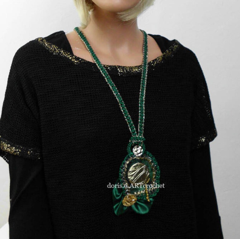 May include: A green and gold crocheted necklace with a large, ornate pendant featuring a silver mirror. The pendant is surrounded by green and gold crocheted flowers.