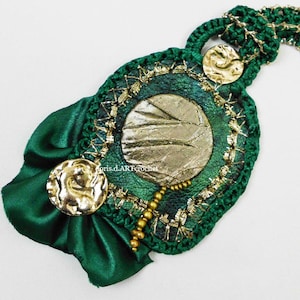 May include: A green crocheted necklace with gold accents and a green satin bow. The necklace features a round pendant with a gold and green design.