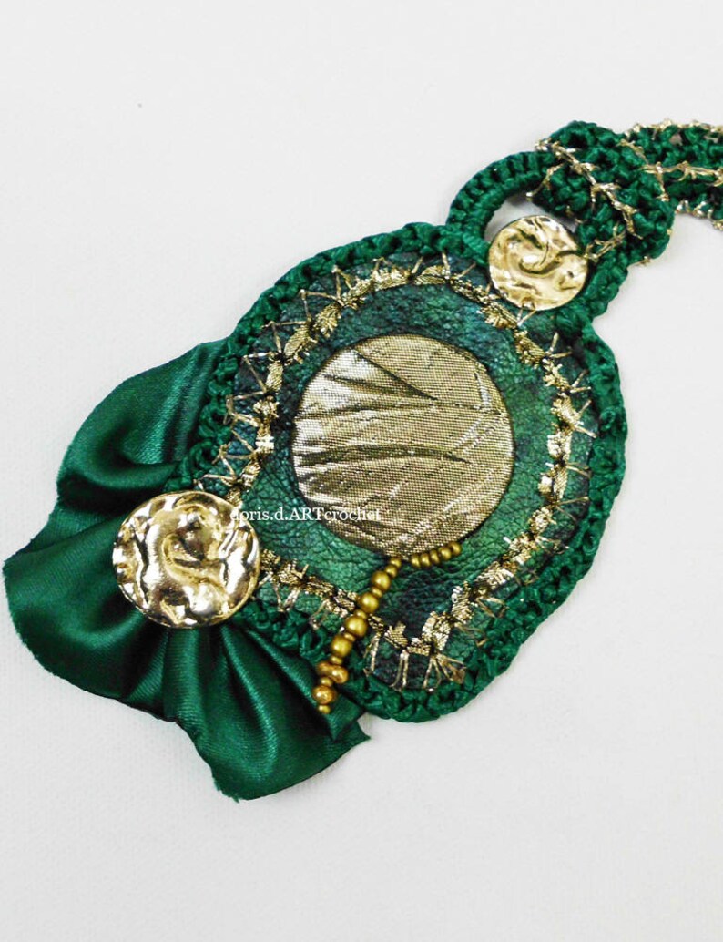 May include: A green crocheted necklace with gold accents and a green satin bow. The necklace features a round pendant with a gold and green design.