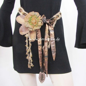 May include: A brown and gold fabric belt with a large flower embellishment and long, flowing fabric tails. The belt is tied in a bow at the front and has a website address, "www.dorisohse.de", printed on the fabric.