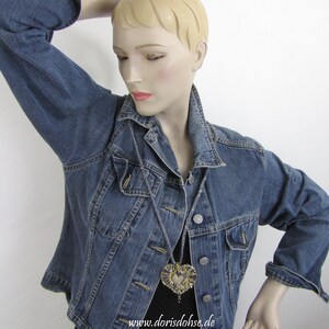 May include: A denim jacket with a heart-shaped pendant necklace. The pendant is made of gold and purple fabric and has a beaded fringe. The necklace is hanging on a mannequin wearing the jacket. The text "www.dorisdohse.de" is visible at the bottom of the image.