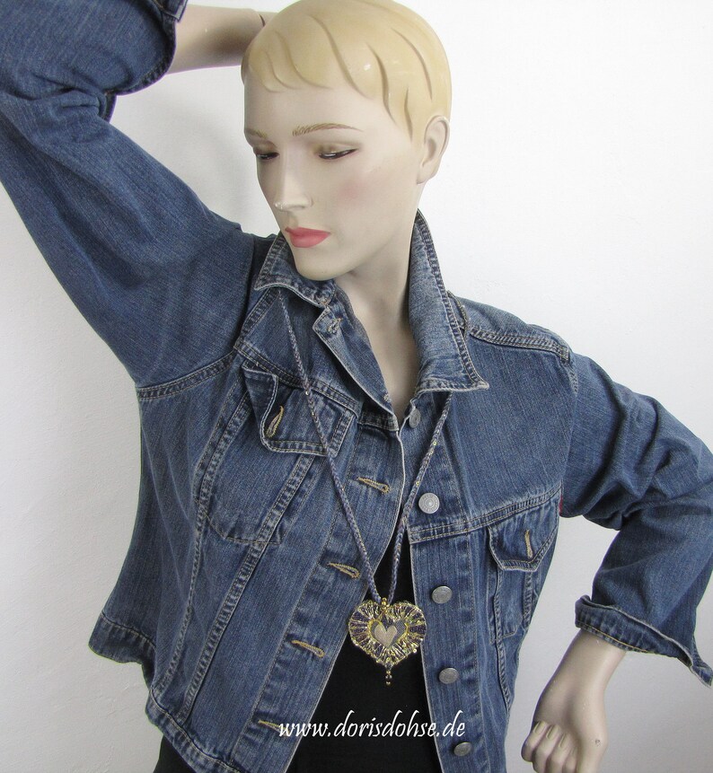 May include: A denim jacket with a heart-shaped pendant necklace. The pendant is made of gold and purple fabric and has a beaded fringe. The necklace is hanging on a mannequin wearing the jacket. The text "www.dorisdohse.de" is visible at the bottom of the image.