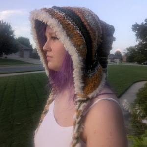 May include: A crochet hood featuring a multi-colored striped pattern in shades of mustard yellow, dark green, black, and white. The hood is trimmed with a thick, fuzzy white faux fur border and includes two long, braided ties hanging down the front.