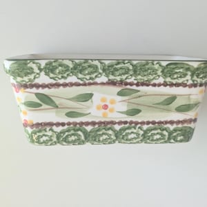 May include: A rectangular ceramic loaf pan featuring a hand-painted pattern of green scalloped borders, brown dotted lines, and a central band decorated with green leaves and small yellow flowers with red centers on a cream-colored background.