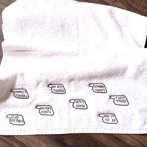 May include: White bath towel with embroidered black text labels. The labels include phrases like "Fawlty Tower", "Watery Towels", and "Fatty Owls". The towel is folded and lies on a wooden surface.