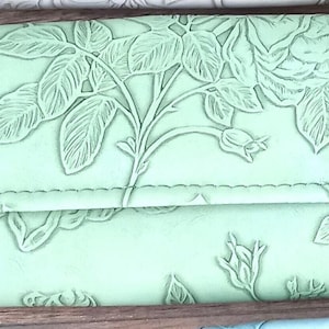May include: A pale green leather wallet featuring an embossed floral rose pattern. The wallet has a flap closure with two gold-coloured snap fasteners. The design includes detailed leaves, stems, and rose buds, offering a tactile and visual appeal.