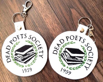 Può includere: Due portachiavi rotondi bianchi con la scritta "DEAD POETS SOCIETY" in nero, che circondano una pila di libri. L'anno "1959" è sotto i libri. Il disegno è circondato da una ghirlanda verde. Ogni portachiavi ha un fermaglio argentato.