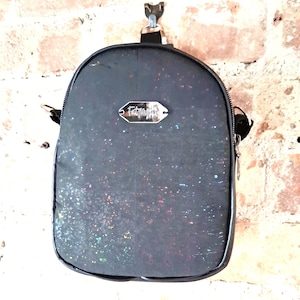 May include: A small, black backpack with a speckled, iridescent design. The bag has a silver tag with the text "Fabriagens". The backpack is hanging against a brick wall.