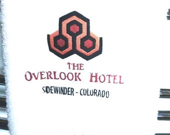 Könnte beinhalten: Weißes Handtuch mit dem Text "THE OVERLOOK HOTEL" in Burgunderrot und Schwarz. Das Handtuch hat ein geometrisches Design in Rot, Schwarz und Orange. Der Text "SIDEWINDER - COLORADO" ist ebenfalls sichtbar.
