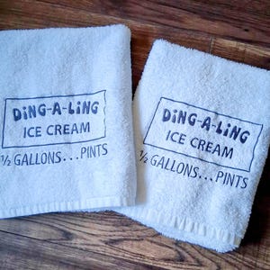 May include: Two white towels with blue embroidery. The towels have the text "DING-A-LING ICE CREAM 1/2 GALLONS...PINTS" on them.