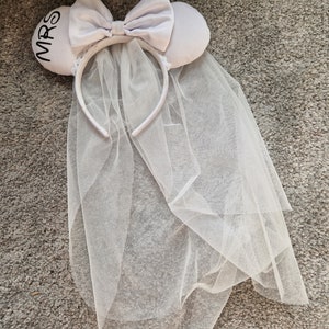May include: White bridal headband with a white tulle veil and a white satin bow. The headband has a white felt ear with the word "MRS" embroidered in black.