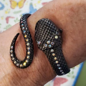 May include: A black snake bracelet with iridescent rhinestones. The bracelet wraps around the wrist, with the snake's head and tail forming the ends. The snake's body is covered in a textured pattern.