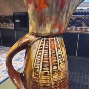 May include: A ceramic pitcher featuring a flared rim and a curved handle. The body displays a textured, grid-like pattern in shades of brown, orange, and cream. The glaze has a crackled finish with blended colors throughout the entire surface.
