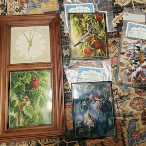 May include: A wooden framed clock with a cream-colored clock face and Roman numerals. Below the clock is a framed picture of two birds in a green grape vine. There are also four framed pictures of birds and certificates of authenticity.