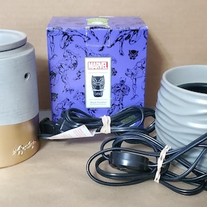 May include: Two gray ceramic wax warmers with black cords and plugs. One warmer has a gold band around the top and the other has a wavy design. The box for the warmer has a purple background with a black and white image of Black Panther on it. The box says "Marvel Black Panther Wax Warmer".