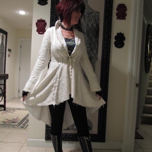 May include: A white, asymmetrical, long-sleeved jacket with a tie at the waist. The jacket is worn over black leggings and black boots with laces.