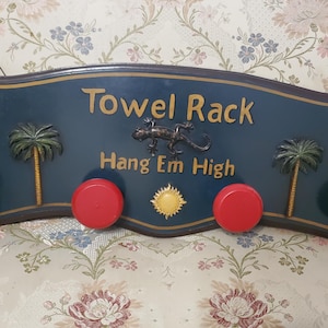 May include: A decorative towel rack with a dark blue background, featuring the words "Towel Rack" and "Hang Em High". It includes red knobs, parrots, palm trees, and a lizard. The sun is also depicted.