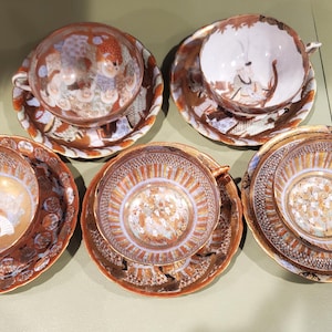 May include: Five antique teacups and saucers. The cups and saucers have a detailed design with orange, brown, and gold accents. The teacups have small handles. The saucers have a scalloped edge. The teacups and saucers are arranged on a light green surface.