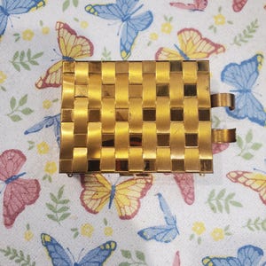 May include: A gold-toned, rectangular compact with a woven pattern. The compact has two curved metal clasps. The background is a white cloth with a pattern of colorful butterflies and small yellow flowers.