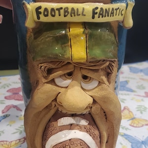 May include: A ceramic mug with a sculpted face design, featuring a football in the mouth. The mug is painted in shades of blue, green, yellow, and brown, with the words "FOOTBALL FANATIC" on a banner.