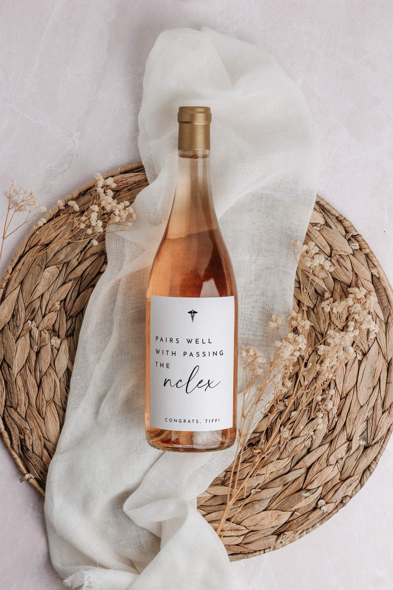 May include: A bottle of ros&eacute; wine with a gold cap and a white label that reads "Pairs Well With Passing The Nclex". The bottle is on a woven placemat with a white cloth and dried flowers.
