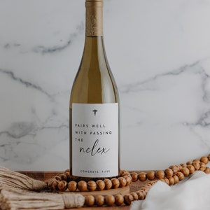 May include: A bottle of white wine with a custom label that reads "Pairs well with passing the NCLEX." The bottle is on a wooden tray with wooden beads and a white cloth. The bottle has a gold foil top.