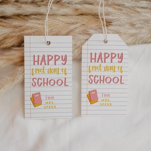 May include: Two white paper tags with the text "HAPPY first day of SCHOOL" in pink and yellow. A small pink book with yellow pages is at the bottom. The tags are lined like notebook paper and say "From MRS. SPEER".