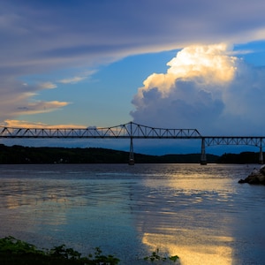 Puede incluir: Un puente cruza un río con una gran nube blanca cumulus en el cielo. El agua refleja el cielo azul y la nube blanca. Una pequeña boya verde y blanca se encuentra en una pequeña isla en el río.