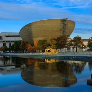 Puede incluir: Un gran edificio redondo de hormigón con un andamio en el lateral. El edificio se refleja en un estanque de agua tranquila. Hay árboles y una escultura dorada en primer plano.