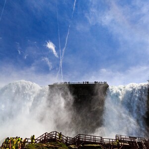May include: A view of Niagara Falls from the observation deck, with a blue sky and white clouds in the background. People are standing on the deck, looking out at the falls.