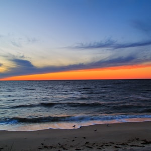 May include: A scenic view of a sandy beach with a calm ocean and a vibrant orange sunset sky. The sky is a mix of blue, orange, and pink hues, with wispy clouds.