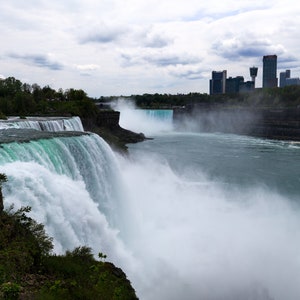 May include: A panoramic view of Niagara Falls, with the American Falls on the left and the Canadian Falls on the right. The falls are surrounded by lush green foliage and a cityscape in the background.
