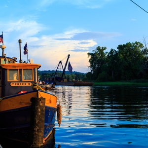 May include: A blue and yellow tugboat named "Tender 3" is docked at a pier. The boat is flying an American flag and has a rope tied to a wooden post. The boat is in a calm body of water with a green grassy shore in the background.