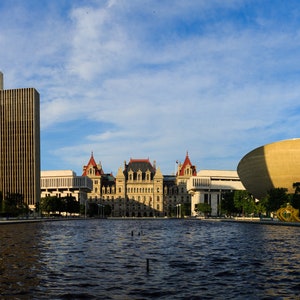 Puede incluir: Una vista del edificio del Capitolio del Estado de Nueva York en Albany, Nueva York. El edificio es una gran estructura blanca con una cúpula y una torre del reloj. En primer plano, hay un estanque reflectante con una gran estructura redonda dorada en el fondo.