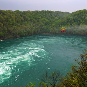 May include: A red and yellow gondola cable car carrying passengers over a swirling, turquoise river surrounded by lush green trees.