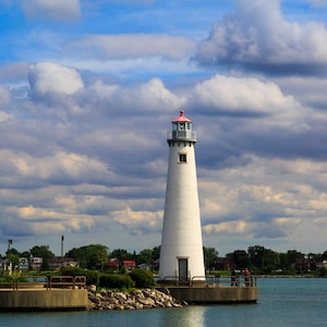 May include: A white lighthouse with a red roof stands on a rocky pier, overlooking a body of water. The sky is a bright blue with fluffy white clouds.