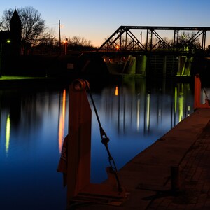 May include: A view of a canal at night with a bridge and a building in the background. The water is reflecting the lights from the bridge and the building. There are two metal posts on the right side of the image.