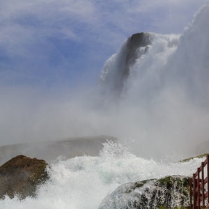 May include: A view of Niagara Falls from a viewing platform. The water is cascading over the edge of the falls and creating a large cloud of mist. Two people are standing on the platform, looking out at the falls.