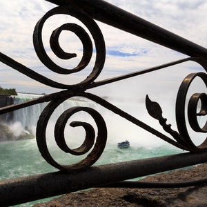 May include: A close-up view of Niagara Falls through an ornate iron railing. The railing is made of black metal with intricate swirls and designs. The falls are in the background, with a boat in the water.