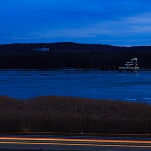 Puede incluir: Una fotografía de larga exposición de un lago azul oscuro con un pequeño edificio iluminado de blanco en la distancia. El lago está congelado y el agua está cubierta de hielo. El edificio está iluminado con luces blancas y está rodeado de árboles. El cielo es azul oscuro y el horizonte es visible en la distancia. Hay hierbas altas en primer plano y una carretera con faros de coche en la parte inferior de la imagen.