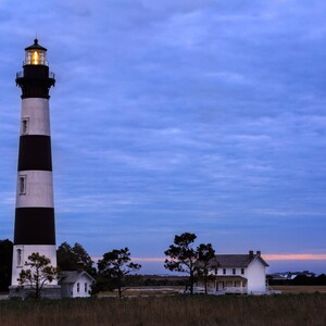 May include: A black and white striped lighthouse with a light on top, against a blue sky with white clouds. A white house with a porch is in the background.
