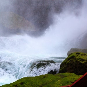 May include: A close-up view of a waterfall with a large amount of mist and spray. The water is crashing over rocks and creating a powerful, white foamy wave. The rocks are covered in green moss. A red wooden structure is visible in the foreground.