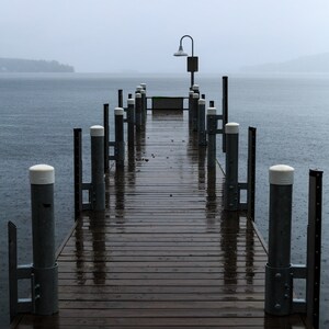 May include: A wooden dock extends out into a lake on a foggy day. The dock is wet from rain and has metal railings with white caps. A single lamp post stands at the end of the dock.