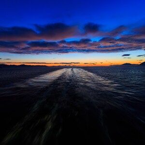May include: A view from the back of a boat as it sails through the water at sunset. The sky is a vibrant mix of blue, orange, and pink, with clouds reflecting the colors. The wake of the boat is visible in the water.