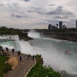 May include: An aerial view of Niagara Falls with a boat in the water and a city skyline in the background. The falls are surrounded by lush green trees and a paved walkway with people walking along it.