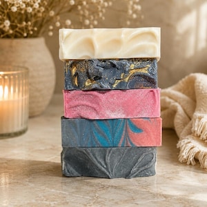 May include: A vertical stack of five rectangular soap bars. The top bar is cream-colored, followed by a dark gray bar with gold swirls, a bright pink bar, a blue and red marbled bar, and a bottom bar in solid dark gray with a leaf-like pattern.